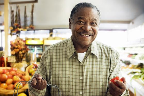 A man smiling in a grocery store, holding a tomato in one hand and a shopping basket in the other hand